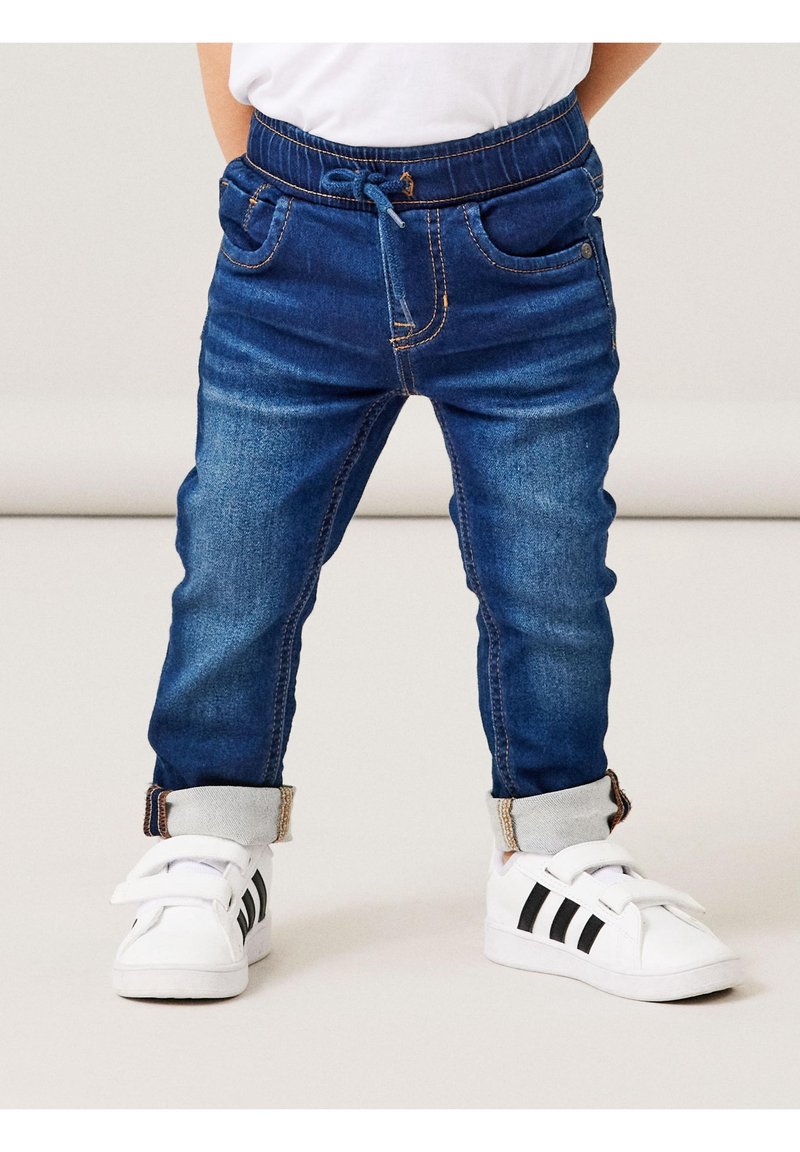 Enfant portant un jean jogger en denim bleu foncé retroussé et des baskets blanches avec des rayures noires, debout sur un sol clair.