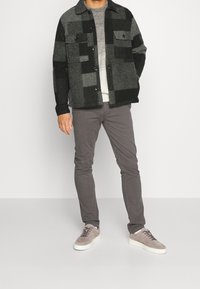 Hombre con chaqueta a cuadros en negro y gris, suéter gris, pantalones grises y zapatillas beige de pie frente a un fondo liso.