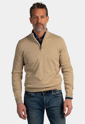 Man met zilvergrijs haar en baard, gekleed in een beige trui met een rits tot de hals over een donkerblauw overhemd en een blauwe spijkerbroek, staand tegen een witte achtergrond.