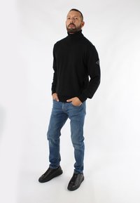 Maglione nero a collo alto, slim fit, abbinato a jeans in denim blu e sneakers nere. Presenta una toppa con logo sulla manica del maglione.