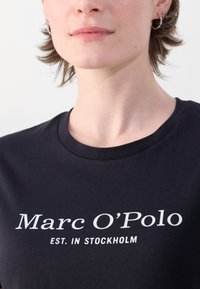 Juodas marškinėliai su apykakle ir trumpomis rankovėmis, turintys baltą tekstą, kurioje parašyta "Marc O’Polo EST. IN STOCKHOLM." Lygus paviršius.