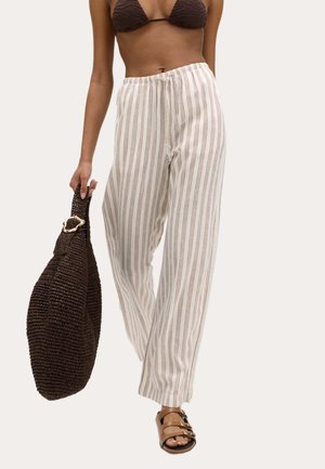 Kobieta ubrana w brązowy top bikini i białe spodnie z brązowymi pionowymi paskami, trzyma dużą brązową plecioną torbę i nosi brązowe sandały.