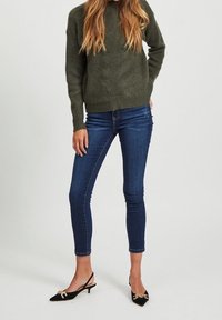 Pull en maille côtelée vert olive, Jean slim-fit bleu marine, et escarpins noirs à bout pointu avec accents en métal doré.