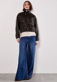 Dunkelbraune Kunstleder-Cropped-Jacke mit hohem Kragen, zwei vorderen Taschen, elastischem Bund und weit geschnittenen blauen Jeans.