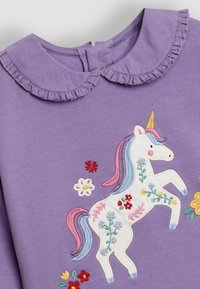 Prenda morada con cuello con volantes, que presenta un diseño de unicornio bordado en blanco, con acentos florales coloridos.