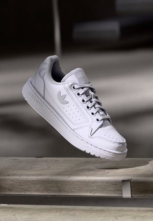 Weißer Adidas-Sneaker mit Schnürsenkeln, der über einer Betonbank schwebt, und zeigt das Adidas-Logo an der Seite und auf der Zunge.