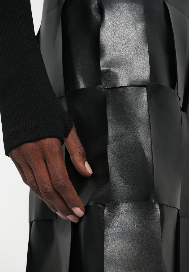 Donkere hand met naakte nagellak rustend tegen een zwart synthetisch leren patchwork rok en een zwarte geribbelde mouw.