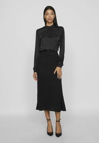 Svart långärmad blus med samlad halsringning, i kombination med en svart räfflad midiklänning. Modellen bär dessutom svarta spetsiga klackskor.