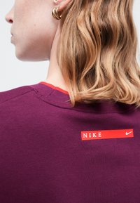 Primo piano di una persona con capelli biondi che indossa una maglietta Nike viola con un'etichetta rossa sul retro vicino alla scollatura, con il nome e il logo del marchio.