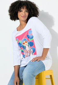 Witte lange mouwen top met een kleurrijke grafische print van een vrouw met blauw haar en een zonnebril, gecombineerd met lichtblauwe jeans.