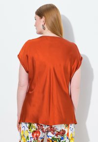 Blusa a maniche corte di colore ruggine con una finitura liscia e satinata. Presenta una vestibilità rilassata e maniche leggermente arrotolate, abbinata a pantaloni con motivo floreale.