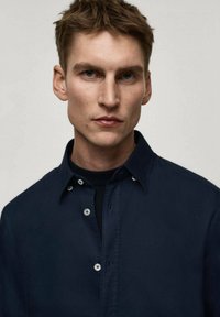 Camicia blu navy con colletto strutturato, tessuto testurizzato e bottoni bianchi, indossata sopra un dolcevita scuro. Dettagli di cuciture sottili visibili.