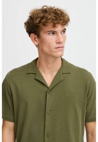 Camisa olive green de manga corta hecha de una tela suave, con un cierre de botones en el frente, cuello abierto y un sutil logo en relieve en el pecho.