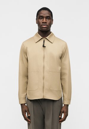 Joven con chaqueta beige con cremallera y grandes bolsillos, y pantalones grises, de pie contra un fondo claro y liso.