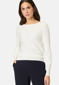 Weißer gerippter Strickpullover mit langen Ärmeln, der horizontale Streifen und einen taillierten Saum aufweist. Getragen mit navyblauen Hosen.
