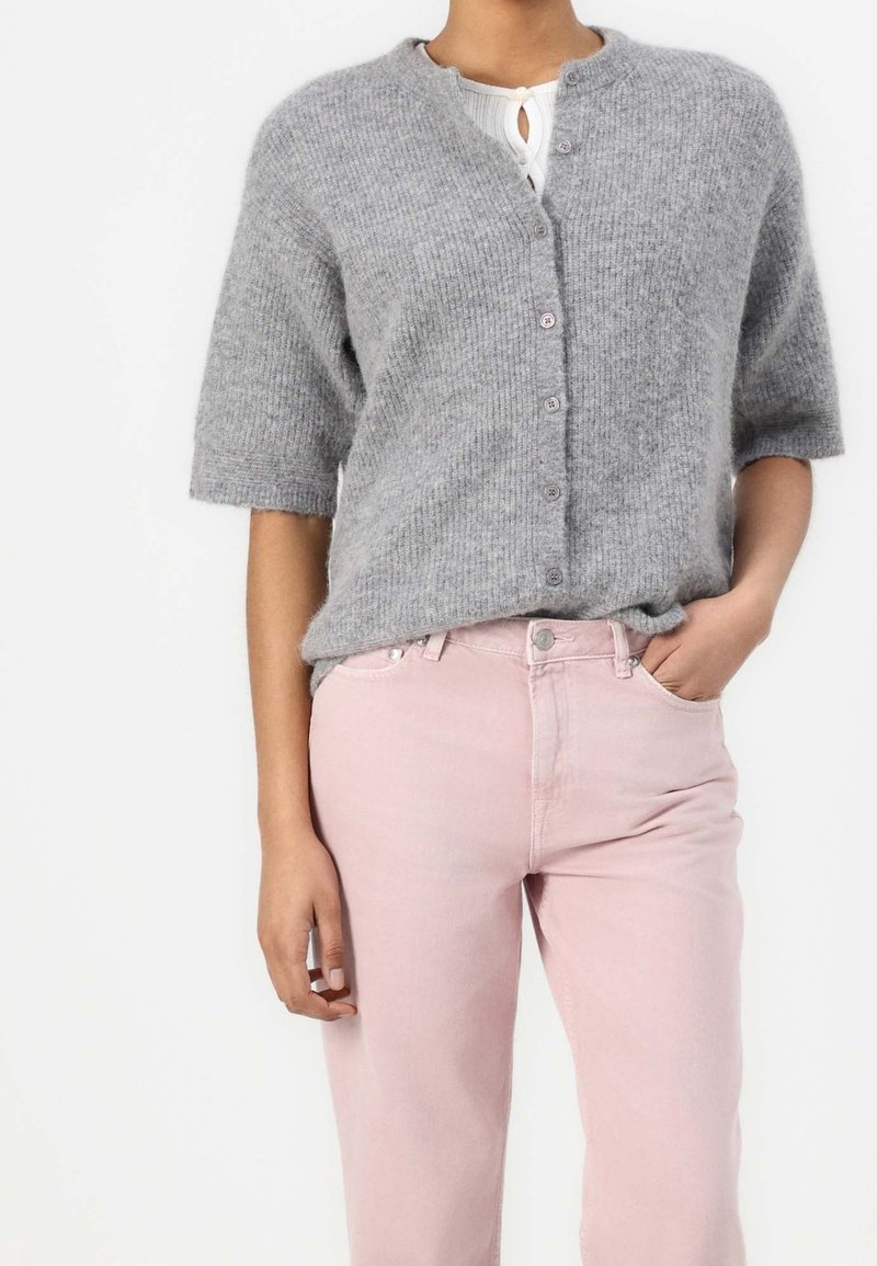 Personne portant un cardigan gris à manches courtes boutonné sur un haut blanc avec un pantalon rose taille haute, une main dans la poche.