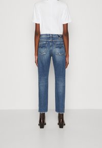 Jeans de mezclilla azul de tiro alto con pierna recta, costuras visibles y dos bolsillos traseros, combinados con una camiseta blanca y botas de tacón oscuro.