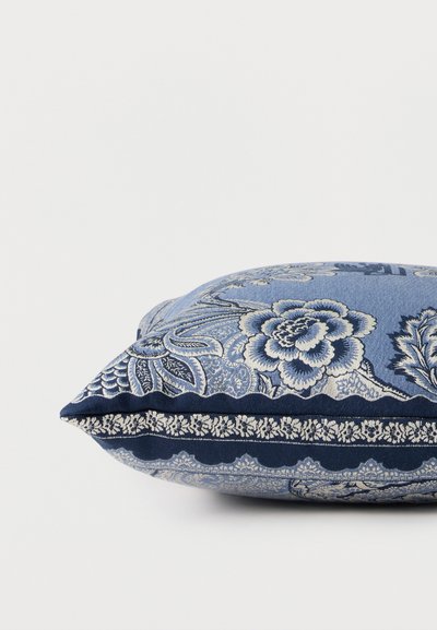 Coussin décoratif bleu avec des motifs floraux et cachemire blancs complexes sur un fond blanc uni.