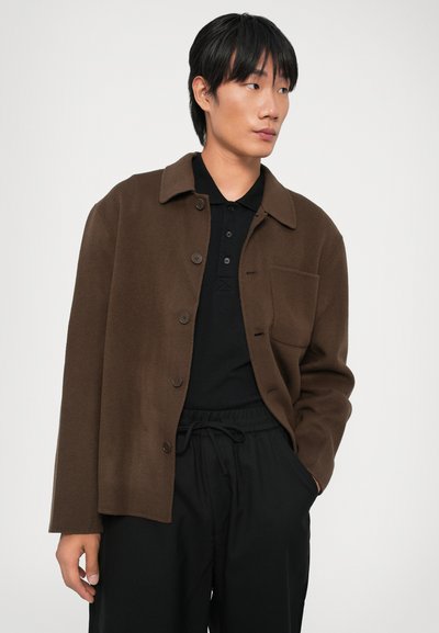 Veste en mélange de laine marron avec un col, fermeture à boutons et poche poitrine, assortie à un polo noir et un pantalon noir à cordon de serrage.