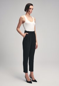 Witte geribbelde tanktop en zwarte op maat gemaakte broek met zijzakken, plooien en een slim fit. Zwarte puntige hakken maken de look compleet.