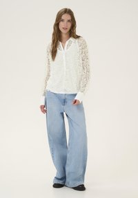 Hvid blondebluse med blomsterprint og krave, parret med lyseblå vidde bukser og sorte sko, der står mod en neutral baggrund.