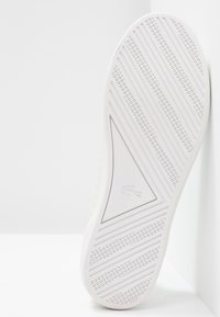 Semelle de basket blanche avec des crampons diagonaux et un logo triangulaire présentant un crocodile au centre sur un fond blanc.