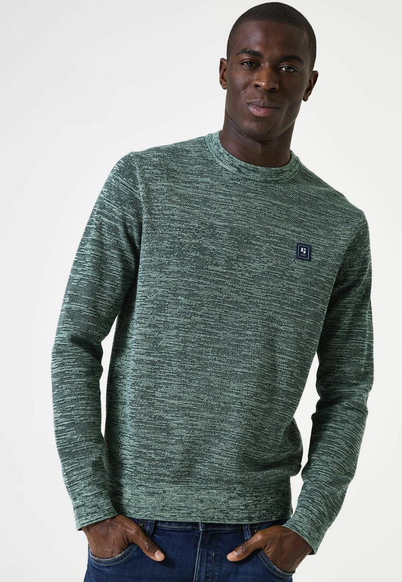 Groene gebreide trui met lange mouwen, ronde halslijn en een klein blauw logo-opgenaaid op de borst. Draag het met blauwe spijkerbroek.