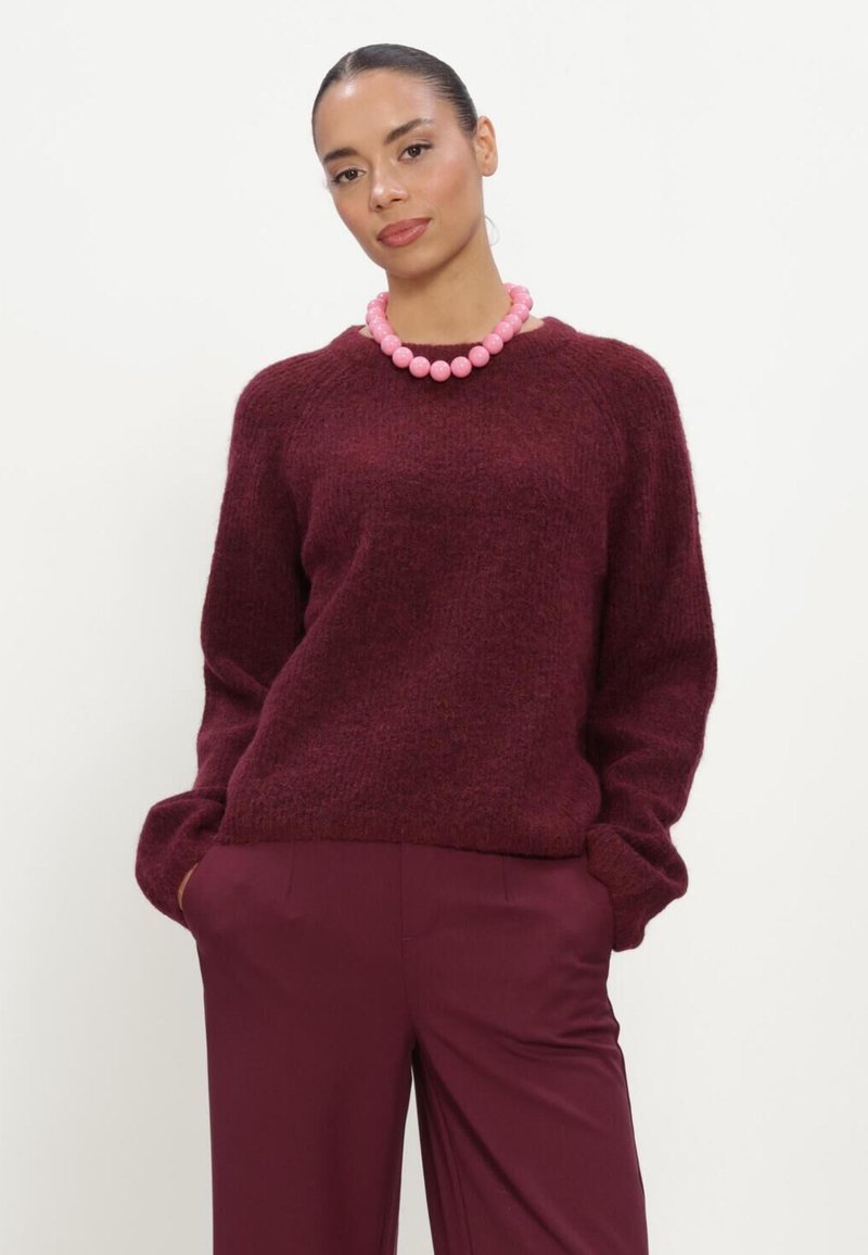 Femme aux cheveux attachés, portant un pull et un pantalon bordeaux avec un gros collier de perles roses, debout les mains dans les poches.
