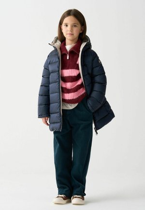 Marineblauer Pufferjacke mit Kapuze über einem gestreiften burgunderroten und rosafarbenen Pullover, kombiniert mit türkisfarbenen Cordhose und braunen Sneakern.