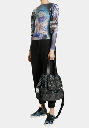 Femme portant un haut transparent à manches longues imprimé avec un motif graphique, un pantalon noir court, des baskets bleues à imprimé léopard, tenant un grand sac à main noir à motif.