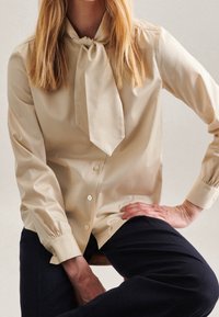 Beige blouse with a tie collar, featuring long sleeves, a button front, and gathered cuffs, made from smooth fabric. Worn with dark pants.