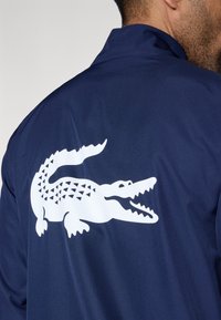 Chaqueta azul marino con acabado texturizado que presenta un gran logo blanco de cocodrilo en la espalda. El cuello es estándar con un diseño liso.