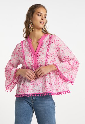 Vrouw die een roze en witte geborduurde blouse met pomponrand en blauwe jeans draagt, poseert tegen een effen witte achtergrond.