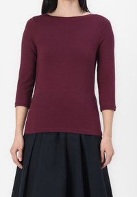 Bordeaux långärmad topp med båtformad halsringning och figurnära design; mjukt bomullsmaterial med en slät textur. Bärs med en svart kjol.