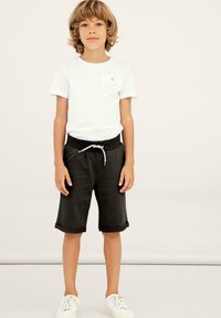 White short-sleeve T-shirt with a small pocket, paired with black knee-length shorts and white sneakers. Soft, casual fabric with a sporty look.