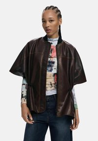 Giacca poncho in pelle marrone con chiusura a zip, colletto a coste e dettagli con coulisse; indossata su una maglietta a maniche lunghe con grafica multicolore.