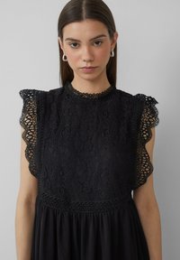 Robe noire avec une superposition en dentelle à motifs floraux, manches à volants et corsage texturé, s'évasant en une jupe douce.