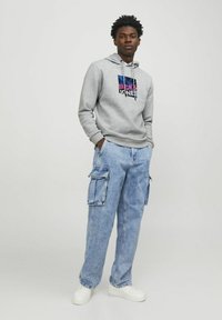 Sudadera gris con un logotipo gráfico en el frente, combinada con pantalones cargo de color azul claro que tienen varios bolsillos y un ajuste relajado, usados con zapatillas blancas.