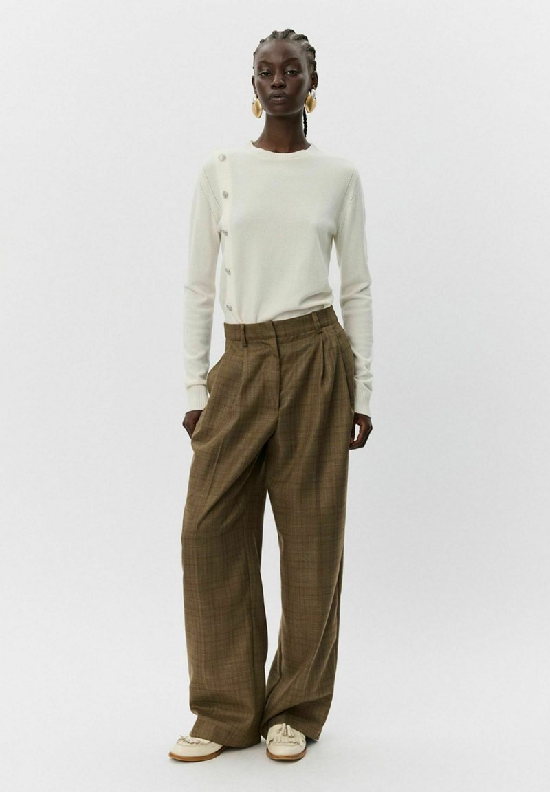 Pull crème clair avec boutons sur le côté, assorti à un pantalon large en plaid marron. Le mannequin porte des chaussures crème, mettant en avant un design texturé.