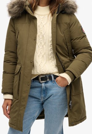 Personne portant un parka vert olive à capuche en fourrure, un pull crème à torsades, un jean bleu clair et une ceinture noire avec une boucle argentée.