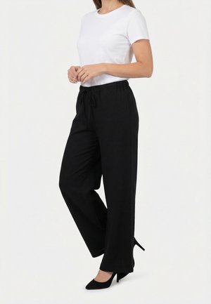 Mujer vestida con una blusa blanca de manga corta, pantalones negros holgados con cordón y zapatos de tacón altos negros, de pie frente a un fondo liso.