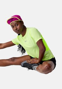 Camiseta atlética amarilla neón de manga corta, combinada con pantalones cortos negros y zapatillas de correr oscuras. El modelo adopta una pose de estiramiento. Una gorra rosa añade color.