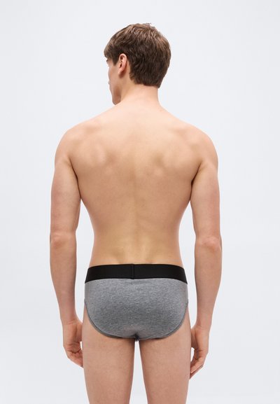 Homme debout de dos, portant un slip gris avec une ceinture noire, sur un fond clair uni.