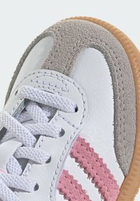 Biały skórzany sneaker z jasnoszarymi i różowymi akcentami, wyposażony w teksturowany panel zamszowy, okrągłe sznurowadła oraz gumową podeszwę.