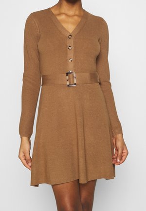 Femme portant une robe en maille côtelée de couleur camel, à manches longues, avec un devant boutonné, un col en V, et une ceinture assortie avec une boucle carrée.