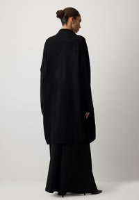 Melna, pārāk liela vilnas mētelis ar šalli apkakli, garām piedurknēm un brīvu piegriezumu, uzvilkts virs melna grīdas garuma svārkiem. Mīksta faktūra.