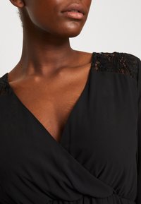 Robe portefeuille noire avec des accents de dentelle sur les épaules, à la texture lisse et au décolleté plongeant. Tissu doux qui tombe élégamment.