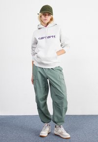 Šedá mikina s kapucí a fialovým logem, zelené cargo kalhoty a šedé a bílé tenisky. Měkké materiály, volný střih a nenáročný design.