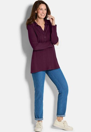 Langemouwen, pruimenblouse met een knoopsluiting bij de kraag en een geplooide voorkant, gedragen met opgerolde blauwe jeans en witte sneakers.