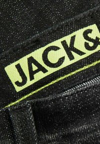 Tessuto in denim nero con un'etichetta gialla visibile con la scritta "JACK &" in lettere nere grassetto, circondata da cuciture a contrasto.